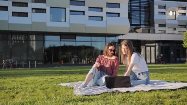 Twee zussen studeren een lezing op afstand op een laptop buiten op de achtergrond van een modern gebouw.. — Stockvideo