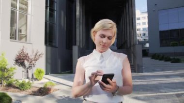 Beyaz tişörtlü güzel ve kendine güvenen sarışın dışarıda akıllı telefon kullanıyor ya da iş arkadaşlarıyla iletişim kuruyor. Takım elbise. Sarışın kız. Başarılı Kadın.