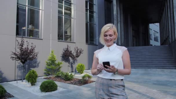 Hermosa y confiada rubia con una camisa blanca lee un mensaje y sonríe al aire libre o se comunica con sus colegas usando el teléfono. Mujer exitosa . — Vídeos de Stock