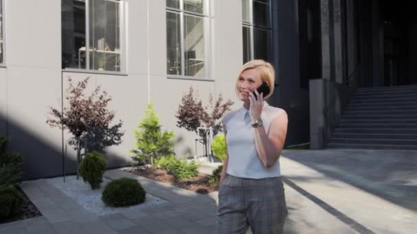 Blonde luxueuse dans une chemise blanche et une belle montre parle sur un téléphone portable et sourit à l'extérieur sur le fond d'un grand bâtiment en verre.Avoir belle coiffure blonde — Video