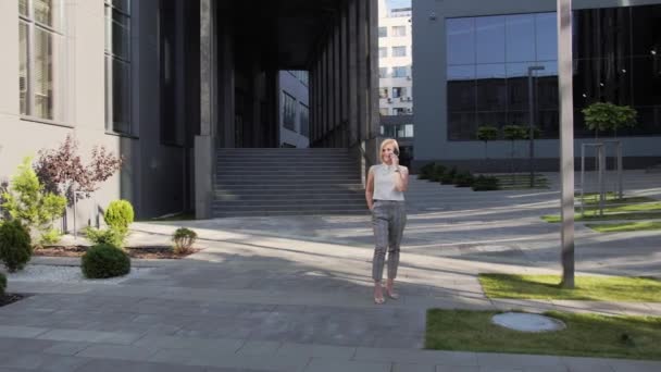 Loira atraente em uma camisa branca e um belo relógio fala em um telefone celular e sorri ao ar livre no fundo de um grande edifício de vidro.Blonde Girl. Mulher de sucesso . — Vídeo de Stock