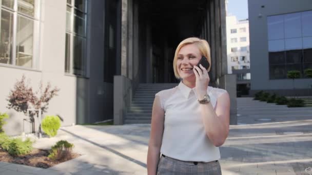 Attraktive Blondine in weißem Hemd und schöner Uhr spricht auf einem Mobiltelefon und lächelt draußen vor dem Hintergrund eines großen Bürogebäudes. Blondes Mädchen. Erfolgreiche Frau. — Stockvideo