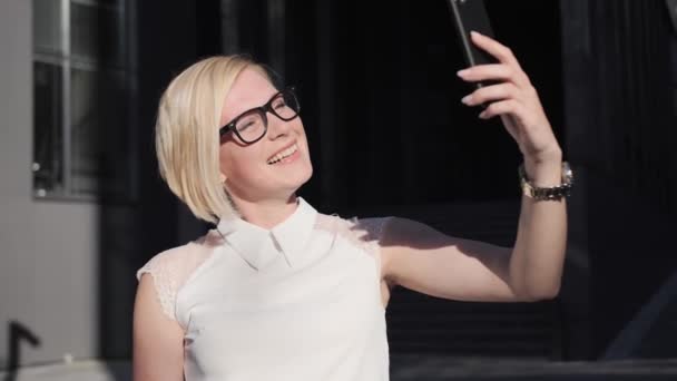 Blond joyeux avec des lunettes parle lors d'un appel vidéo, souriant en utilisant le téléphone sur le fond d'un bâtiment moderne en plein air. Lumière du soleil, blonde d'affaires, le concept du succès. — Video
