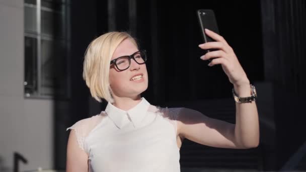 Mujer segura rubia con gafas habla en una videollamada, sonriendo con el teléfono en el fondo de un edificio moderno al aire libre. Luz del sol, rubia de negocios . — Vídeo de stock