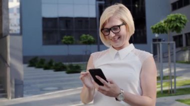 Beyaz tişörtlü çekici sarışın kadın mobil uygulama kullanarak telefonla karşılık veriyor. Kendine güvenen bayan internetten alışveriş yapıyor ve gülümsüyor. İş elbisesi, Sarışın Kız. Başarılı Kadın.