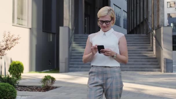 Krásná a sebevědomá blondýna v bílé košili číst sms na mobilním telefonu venku a pak se podívat do kamery nebo chatování s kolegy. — Stock video