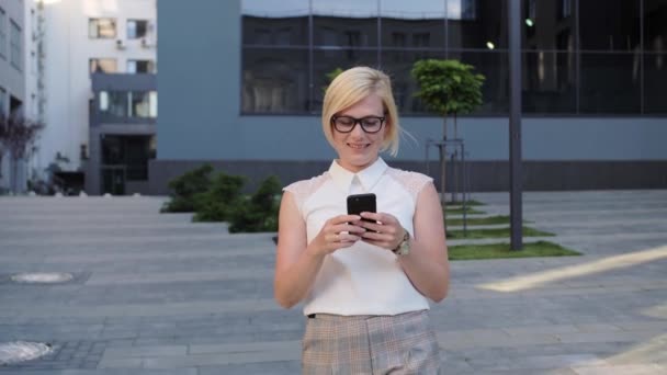 Loira bonita e confiante em uma camisa branca vai e lê a mensagem.Mulher de sucesso. Telemóveis. Aplicações . — Vídeo de Stock