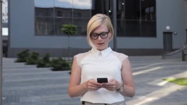 Gözlüklü güzel sarışın dışarı çıkıyor. İş görüşmesi ayarlamak için akıllı telefon kullanıyor. Takım elbise giyiyor, güzel bir saç stili ve şık bir saati var..