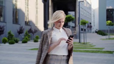 Güzel saç stili olan sarışın iş kadını ofis merkezinin yanında duruyor ve akıllı telefonuna mesajlar yazıyor. Başarılı kadın..