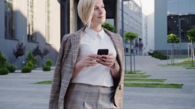 Güzel saç stili olan güzel sarışın iş kadını ofis merkezinin yanında duruyor ve akıllı telefonundan gülümsediği mesajını okuyor. Başarılı kadın..