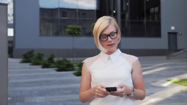 Όμορφη ξανθιά με γυαλιά επιχειρηματική κυρία με τα πόδια σε εξωτερικούς χώρους χρησιμοποιεί ένα smartphone που είναι ντυμένη με ένα επαγγελματικό κοστούμι και έχει όμορφα στυλ μαλλιά και ένα κομψό ρολόι. — Αρχείο Βίντεο