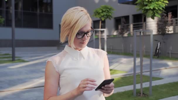 Attraente donna d'affari con gli occhiali va all'aperto utilizza un telefono cellulare per comprare qualcosa che è vestita in un abito da lavoro e ha capelli splendidamente in stile e un orologio alla moda. — Video Stock