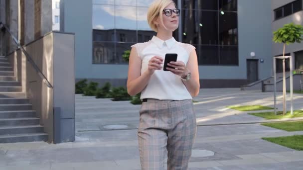 Blondynka bizneswoman odwraca wzrok, a następnie patrzy na telefon komórkowy i przerzuca wiadomości za pomocą smartfona. Stylowe ubrania, okulary, luksusowy wygląd. — Wideo stockowe