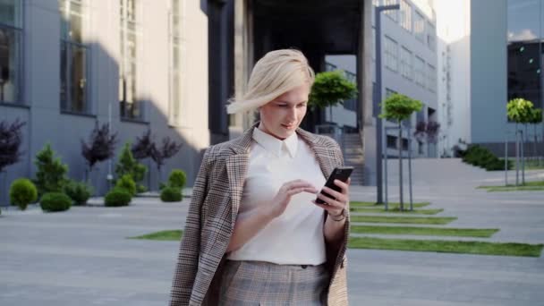 Blonde businesswoman with beautifully styled hair stands near the office center and types messages on her smartphone. Successful woman. — Stock Video