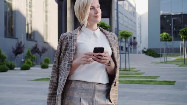 Όμορφη ξανθιά επιχειρηματίας με όμορφα στυλ μαλλιά στέκεται κοντά στο κέντρο του γραφείου και διαβάζει το μήνυμα στο smartphone της χαμογελάει. Επιτυχημένη γυναίκα. — Αρχείο Βίντεο