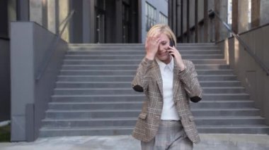 Güzel sarışın iş kadını işten çıkıyor ve cep telefonuyla konuşuyor. Güzel bir iş elbisesi ve güzel bir saç stili giymişti..