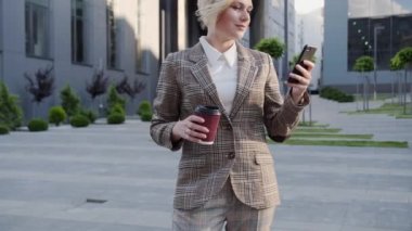 Kahve takımlı güzel sarışın modern bir binanın arka planında telefonunu kullanarak bilet sipariş ediyor. Kendine güvenen, güzel görünümlü bir kadın..