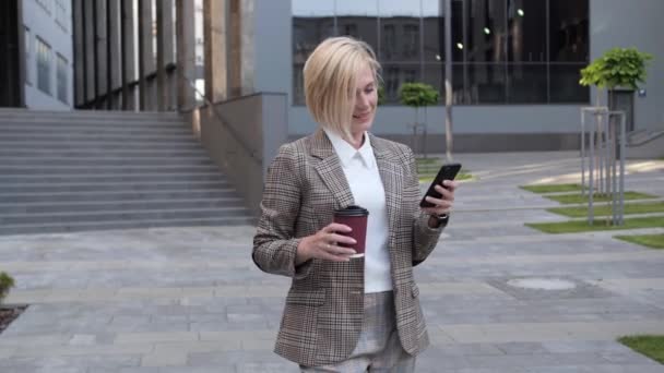 Mulher de negócios loira confiante lê e-mail no telefone celular enquanto bebe café ao ar livre. Aplicações de terno de negócios . — Vídeo de Stock