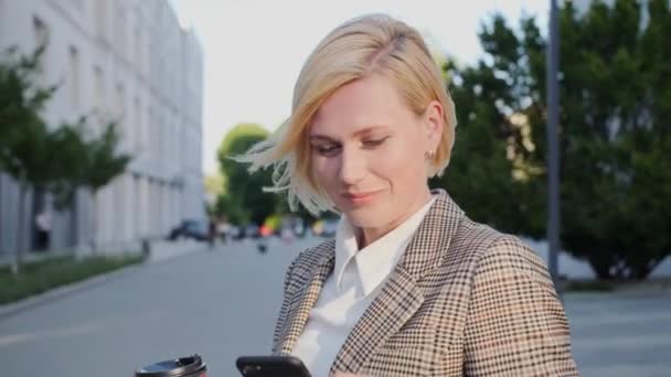 Okouzlující úsměv obchodní blondýny držící telefon a kávu na pozadí moderní budovy.Úspěšná žena. — Stock video