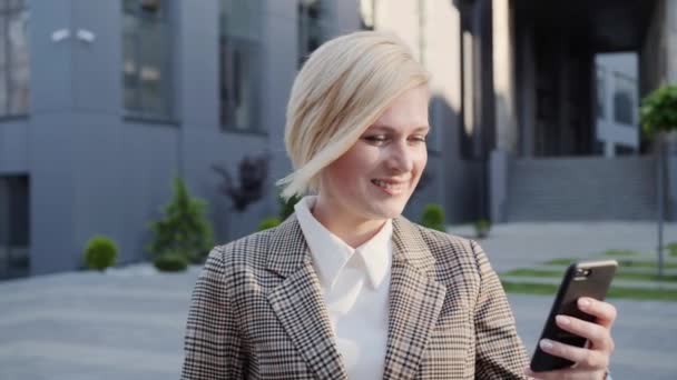 Sorriso affascinante di una bionda d'affari che tiene un telefono che beve caffè sullo sfondo di un edificio moderno.Donna di successo. — Video Stock