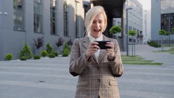 Γοητευτική ξανθιά παίζει ένα παιχνίδι στο τηλέφωνο χρησιμοποιώντας μια εφαρμογή για κινητά στο παρασκήνιο ενός σύγχρονου κτιρίου. Τηλέφωνο, εφαρμογές — Αρχείο Βίντεο