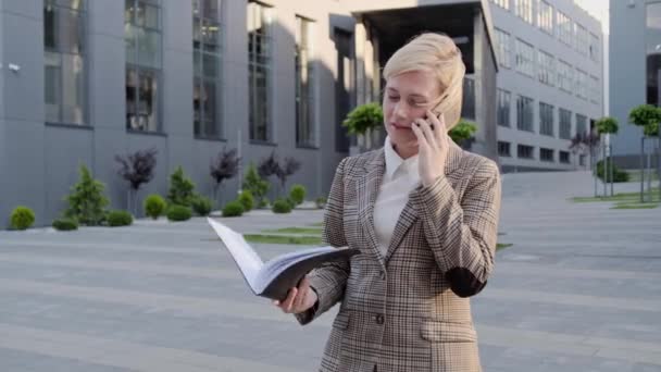 Affascinante bionda tiene un quaderno e parla al telefono all'aperto sullo sfondo del centro ufficio. — Video Stock