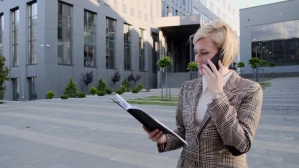 Atraktivní blondýna drží notebook a mluví po telefonu venku na pozadí kancelářského centra. — Stock video