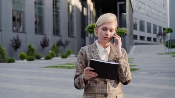 Loira bonita mantém um caderno e fala ao ar livre no telefone no fundo do centro de escritório . — Vídeo de Stock