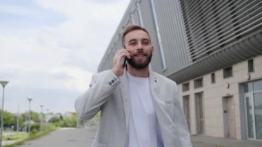 Sakallı genç işadamı, açık havada akıllı telefonunu kullanarak bir iş görüşmesini tartışıyor. Açık renk takım elbise ve beyaz tişört giyiyor..