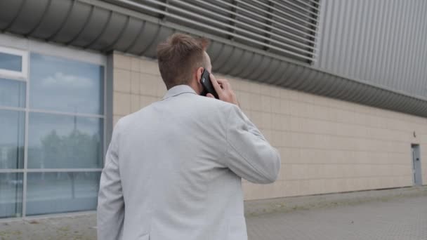 Giovane uomo d'affari con la barba parla di un incontro d'affari utilizzando il suo smartphone all'aperto.Vestito con un abito leggero e t-shirt bianca. Rete globale. — Video Stock