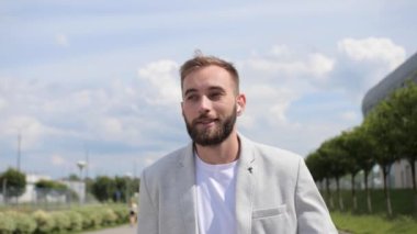 Sakallı genç işadamı modern bir binanın arka planındaki kablosuz kulaklıklardan konuşuyor. Parlak takım elbise, tişört, küresel ağ, kapatın..