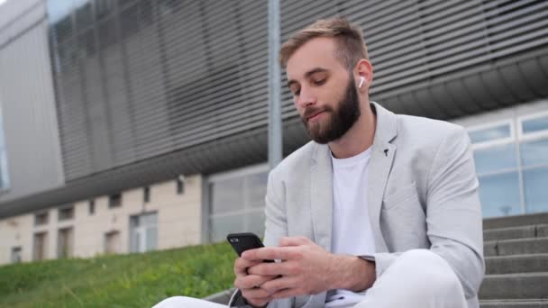 Mladý podnikatel s vousy poslouchá hudbu přes bezdrátová sluchátka na pozadí moderní budovy.Světelný oblek, tričko, globální síť, mobilní aplikace. — Stock video