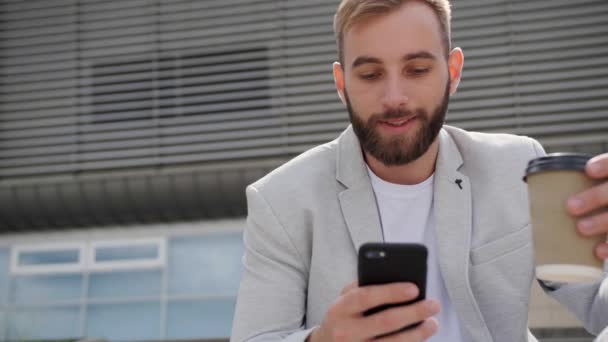Ein junger Geschäftsmann mit Bart sitzt auf der Treppe und blickt auf ein Smartphone, während er vor dem Hintergrund des modernen Gebäudes Kaffee trinkt. Kaffeepausen.Leichter stylischer Anzug, globales Netzwerk. — Stockvideo