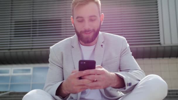 Un giovane uomo d'affari fiducioso con la barba si siede sulle scale e guarda uno smartphone mentre beve caffè sullo sfondo di un edificio moderno. Caffè break.Light abito elegante, rete globale. — Video Stock