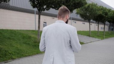 Sakallı ve akıllı telefonlu genç bir bankacı bankaya gider ve elinde kahve tutar. Parlak şık takım elbise, küresel ağ, arkadan para çevrimiçi görünümü.