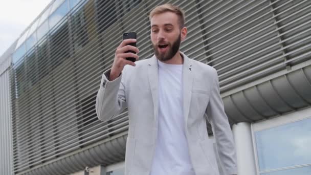 A handsome guy with a beard reacts emotionally to the success of the project, a happy businessman dressed in a stylish light jacket, a mobile application, social networks positive emotion. — Stock Video