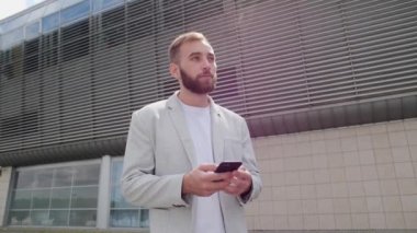 Sakallı ve açık renk ceketli genç adam mobil uygulama kullanıyor ve açık havada duygusal tepki veriyor. Telefondaki oyunlar, uygulamalar, tarayıcılar.