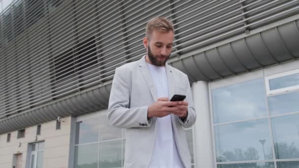 Un jeune homme d'affaires lit un e-mail et se réjouit du succès de son entreprise. Il porte une veste légère et un T-shirt blanc sur le fond du centre de bureau. Personnes qui réussissent, téléphone. — Video