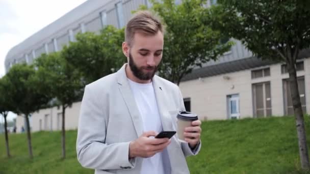 Attraktiver Geschäftsmann mit Bart, der eine Nachricht tippt und einen Kaffee in der Hand hält. In heller Jacke und weißem T-Shirt vor dem Hintergrund eines modernen Gebäudes. — Stockvideo