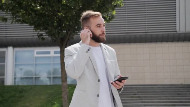 Knappe kerel met een baard praten via draadloze hoofdtelefoon met behulp van mobiele telefoon, app achtergrond modern gebouw licht pak internet. — Stockvideo