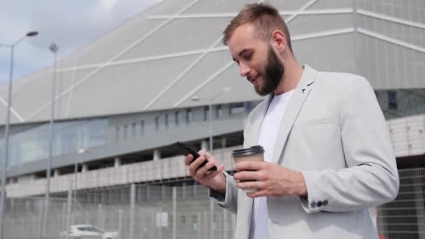 Ein junger selbstbewusster Geschäftsmann mit Bart sitzt auf der Treppe und blickt auf ein Smartphone, während er vor dem Hintergrund des modernen Gebäudes Kaffee trinkt. Kaffeepausen.Leichter stylischer Anzug, globales Netzwerk. — Stockvideo