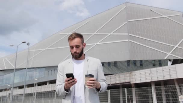 Молодий бізнесмен в легкому костюмі йде з кавою і використовує свій телефон, щоб спілкуватися з колегами на фоні сучасної будівлі, посміхаючись, ділових партнерів . — стокове відео