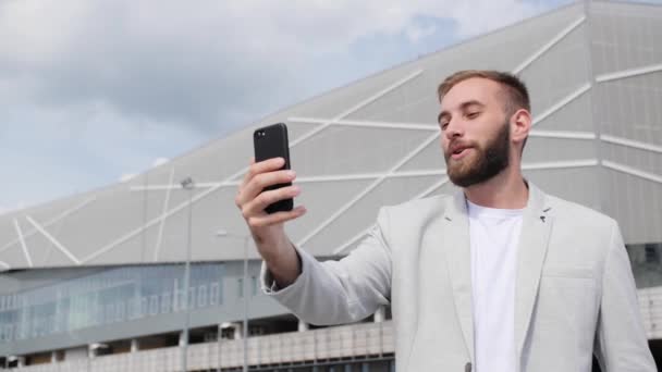 Aantrekkelijke zakenman spreekt op een videogesprek, glimlachend met de telefoon op de achtergrond van een modern gebouw buiten. Zonlicht, het concept van succes. — Stockvideo