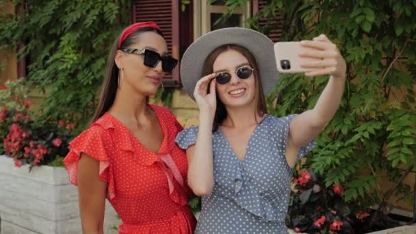 Dua gadis cantik dalam gaun musim panas mengambil selfie di latar belakang rumah yang indah dengan bunga di tengah kota. Gadis itu mengenakan topi dan kacamata hitam. — Stok Video