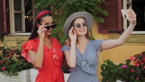 Две лучшие симпатичные сестры в солнцезащитных очках и шляпе веселятся, смеются, делают селфи и смотрят на фотографии на фоне красивого дома с цветами в городе. — стоковое видео