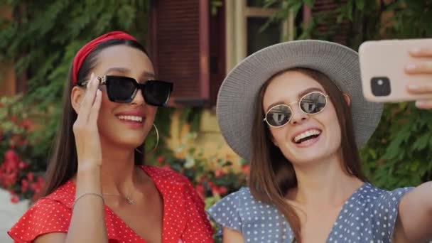 Zwei beste Freunde mit Sonnenbrille und Hut amüsieren sich, lachen, machen Selfies vor dem Hintergrund eines schönen Hauses mit Blumen in der Stadt. — Stockvideo