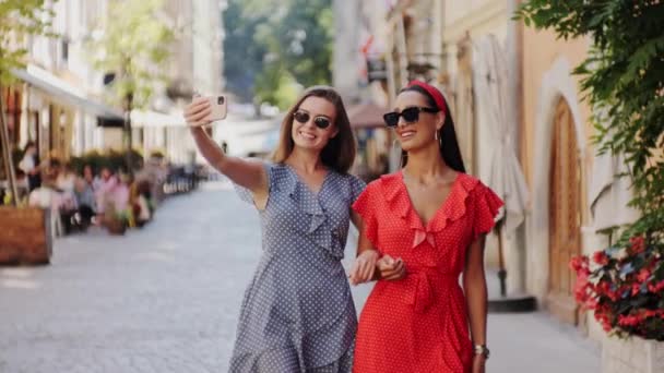 Zwei fröhliche und glückliche attraktive Freundinnen in stylischem Look spazieren durch die Straßen der Stadt. Freundinnen machen Selfies. Telefonfrauen im Freien. — Stockvideo