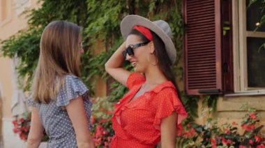 Yazlık elbiseli, güneş gözlüklü, şapkalı, kucaklaşan ve konuşan iki çekici arkadaş şehirde çiçekli güzel bir evin arka planında iyi eğlenceler..