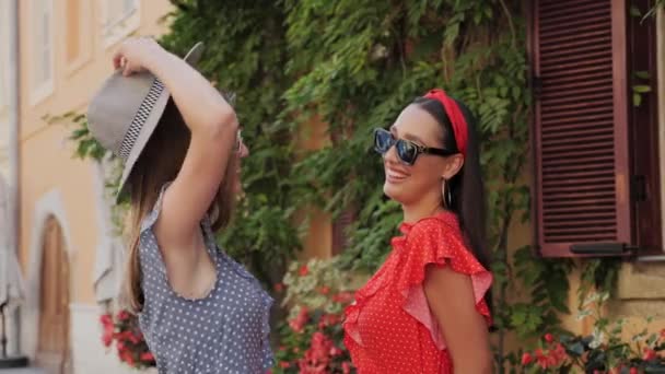 Dos atractivos mejores amigos en vestidos de verano, gafas de sol y sombrero hablando y riendo se divierten en un fondo de una hermosa casa con flores en la ciudad. — Vídeos de Stock