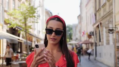 Güneş gözlüklü şık ve güzel bir turist birçok kafenin olduğu şehir caddesinde yürüyor. Kız telefonda konuşuyor..